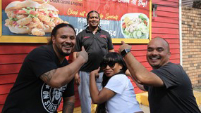 At center, Chef Shermond Esteen, Jr. poses in front of his restaurant as the roadtrippers—from left, Cordero, London, and Hugo—smile and point back at him.